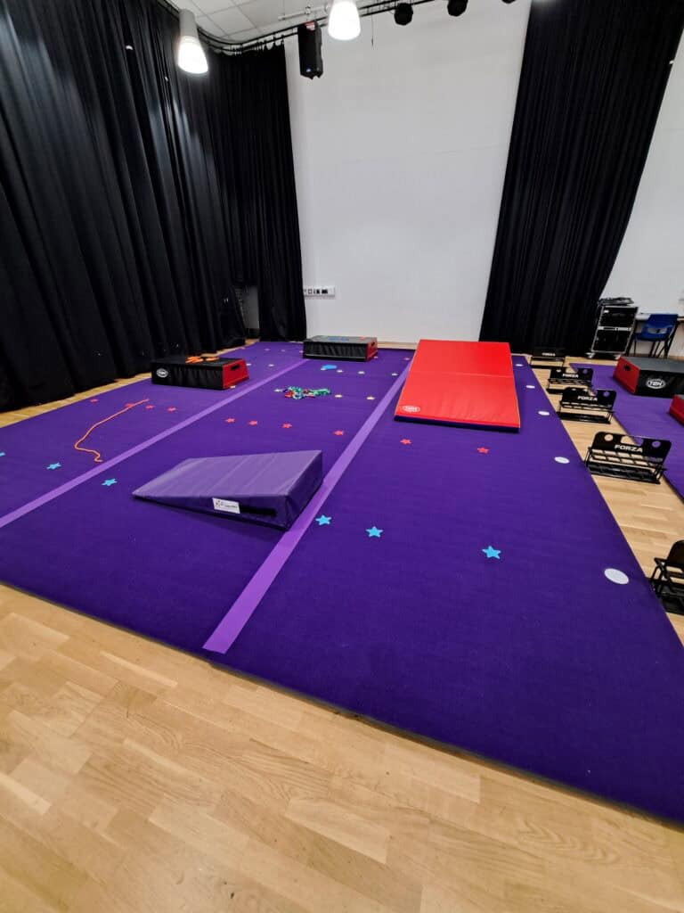 Indoor gymnastics training area with purple mats, foam blocks, and climbing equipment in a studio with black curtains and wooden flooring.