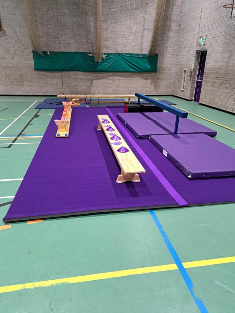 An indoor gymnastics training area with purple mats, balance beams, and training equipment set up on a green sports hall floor.