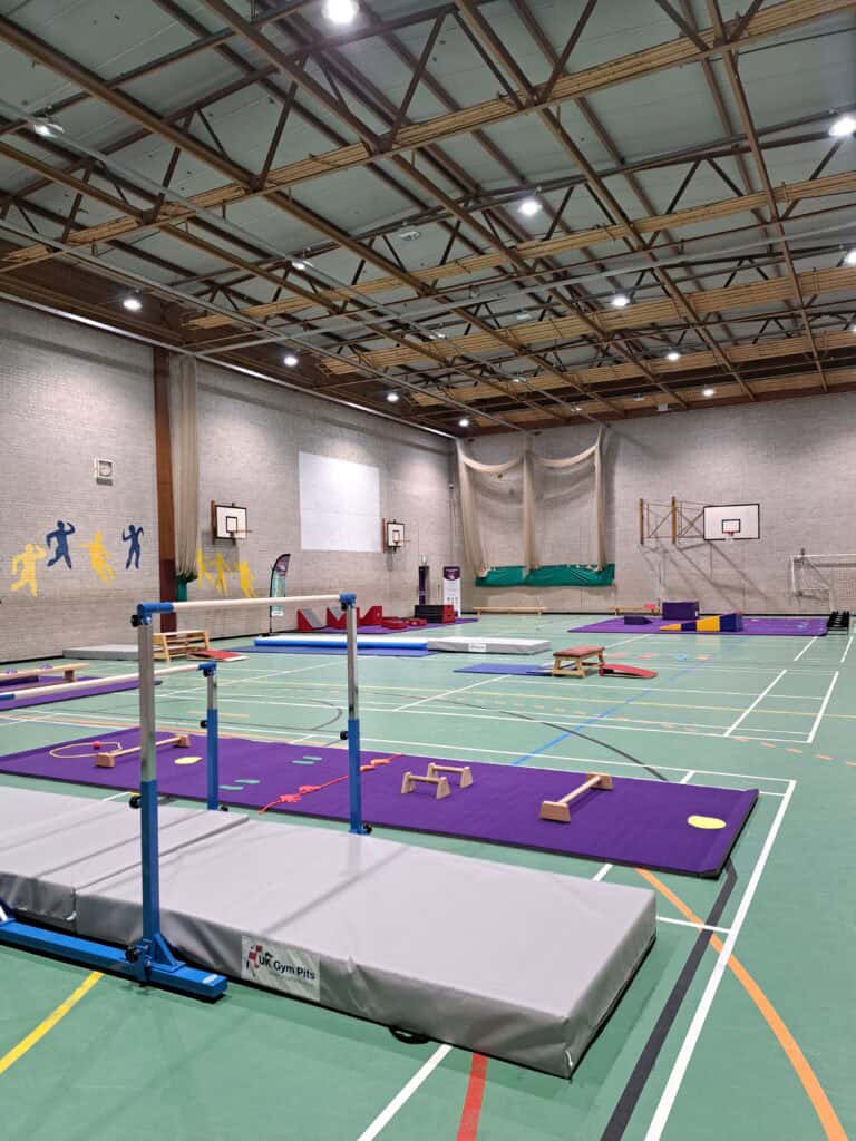 An indoor sports hall equipped for gymnastics training with various mats, bars, and climbing frames, illuminated by ceiling lights and featuring basketball hoops on the walls.