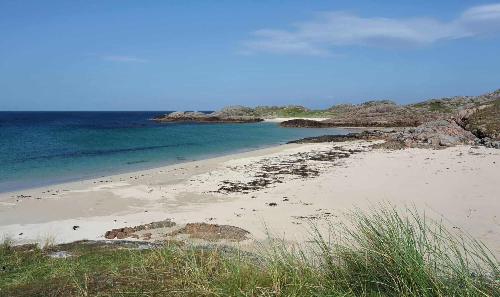 Alt text: Pristine beach with white sand, clear blue waters, and rugged rocks under a partly cloudy sky.