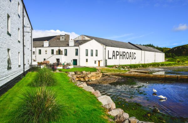 White coastal buildings with "LAPHROAIG" text, lush green grass, and seagulls by the water, capturing the essence of Hebridean adventures.
