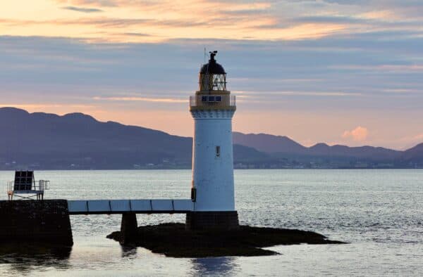 Lighthouse at sunset over calm waters, scenic Scottish coastline with mountain range in the distance.