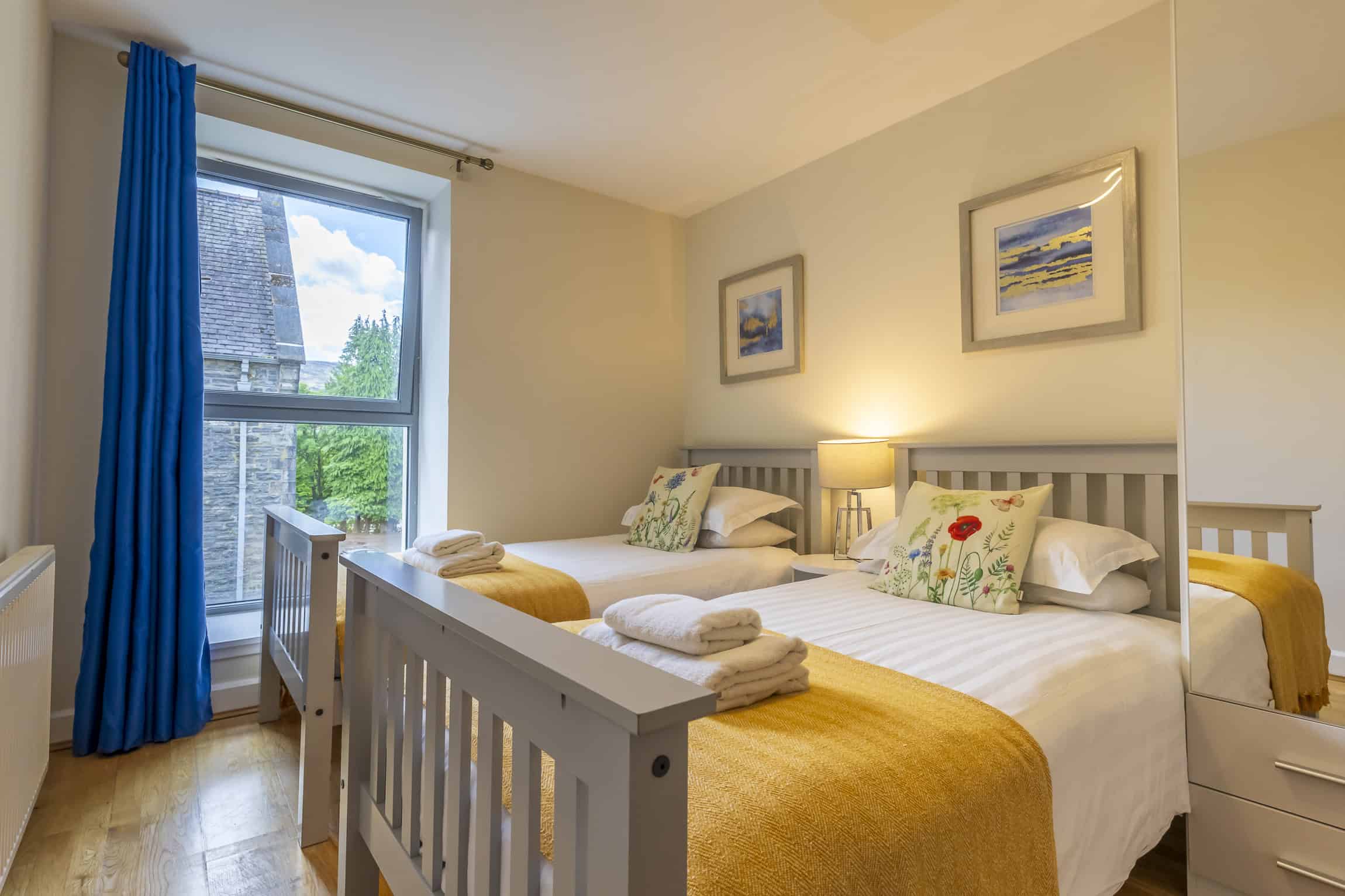 Bright bedroom with twin beds, colourful cushions, and a balcony view of Highland scenery. Perfect f.