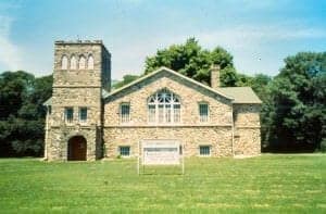 Old stone church building with a tower and surrounding green lawns, part of The History Museum exhibits.