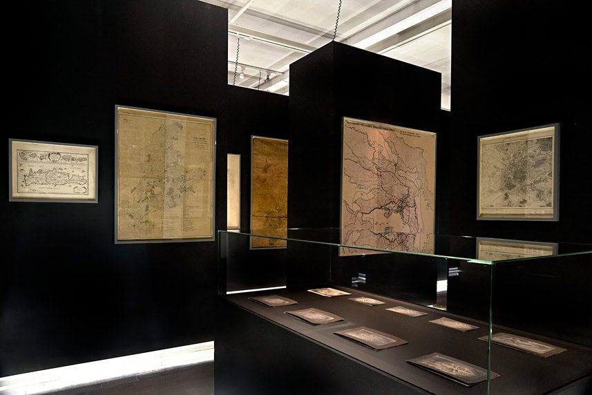 Historical maps displayed in a modern exhibition space with black walls.