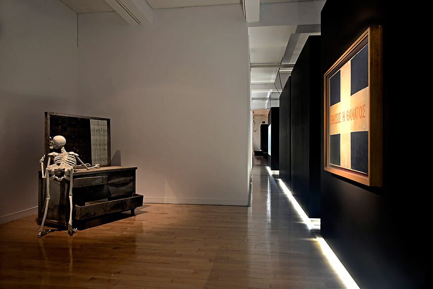 Modern art gallery interior with skeleton sculpture and framed artwork on black wall.