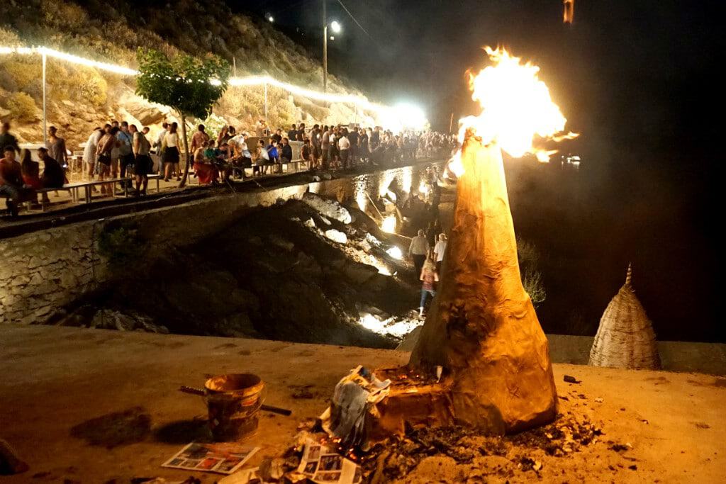Large bonfire at Putiferio festival with crowd and scenic coastline at night.