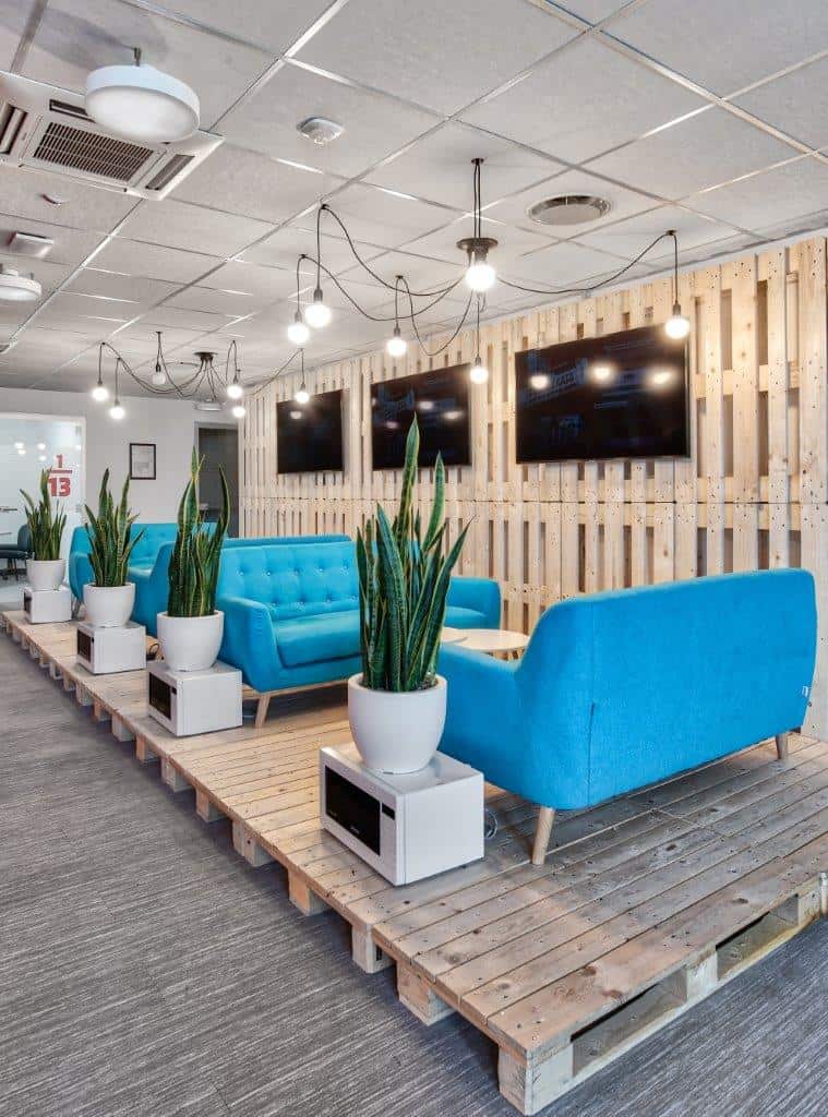 Modern lounge area with blue sofas, potted plants, and wall-mounted TVs.