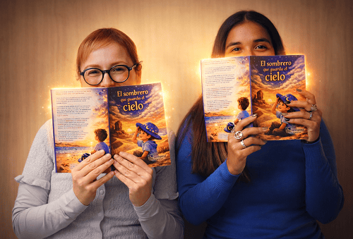 Two women sit side by side, each holding a colorful book titled 'El sombrero que guarda el cielo' up to their faces and smiling behind the pages.
