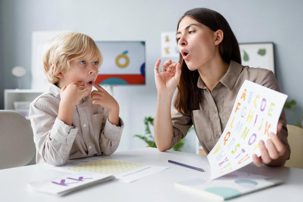 Teacher helps a young boy with letter sounds at a table, holding a colorful alphabet sheet