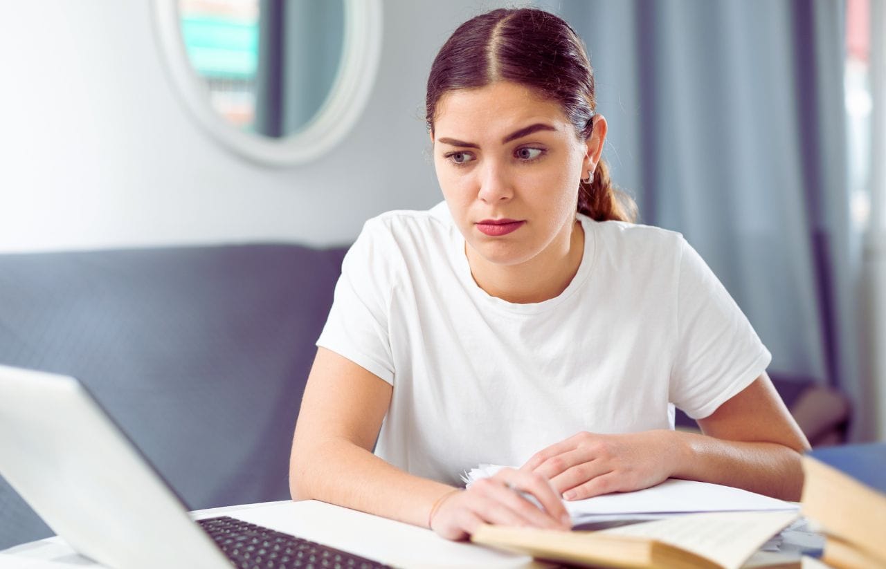 Woman sits at a desk with an open laptop, jotting notes on papers and looking thoughtful.
