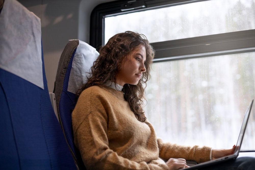 Woman sitting on a bus seat, typing on a laptop by a rain-speckled window.