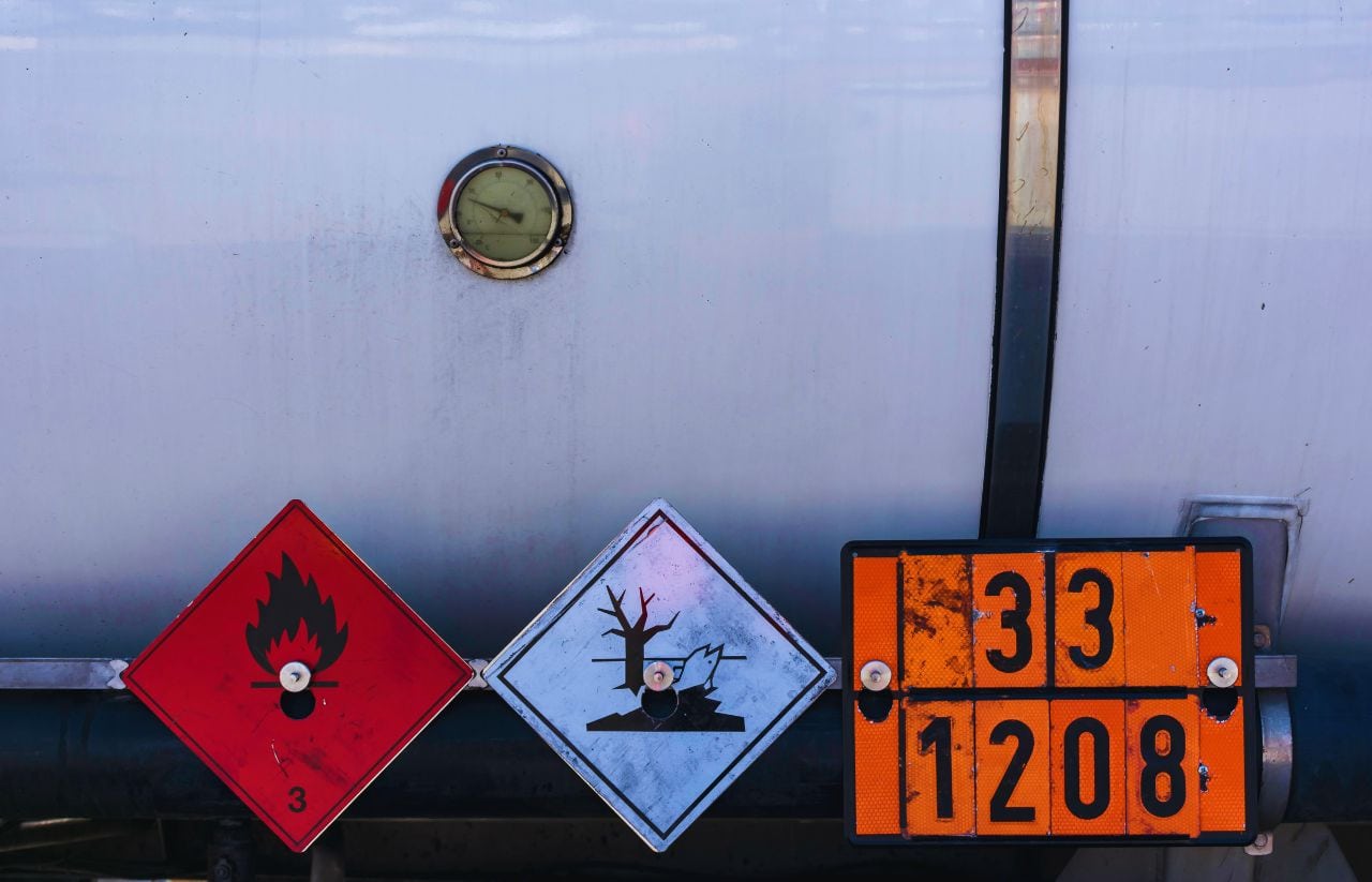 Back of a chemical tanker with three hazard signs: red flammable liquid diamond, blue environmental damage diamond, and an orange class 3/UN placard reading 33 1208