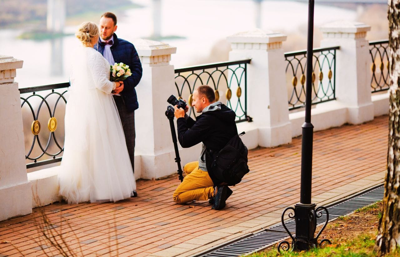 Bride in a white wedding dress and groom in a blue jacket pose by a railing as a photographer shoots them by the waterfront railing.