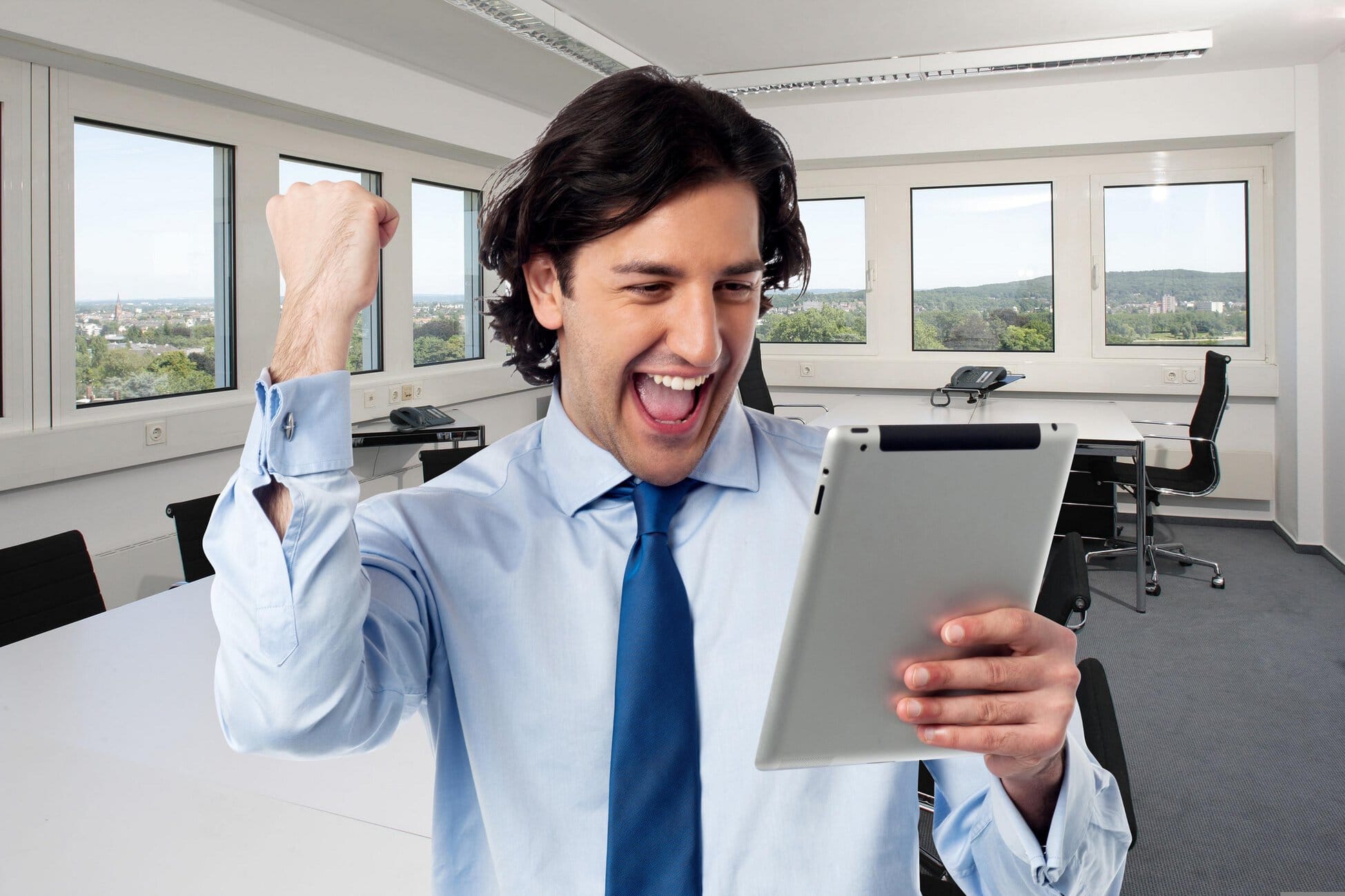 Young man in a blue shirt and tie cheerfully looking at a tablet in a bright office.