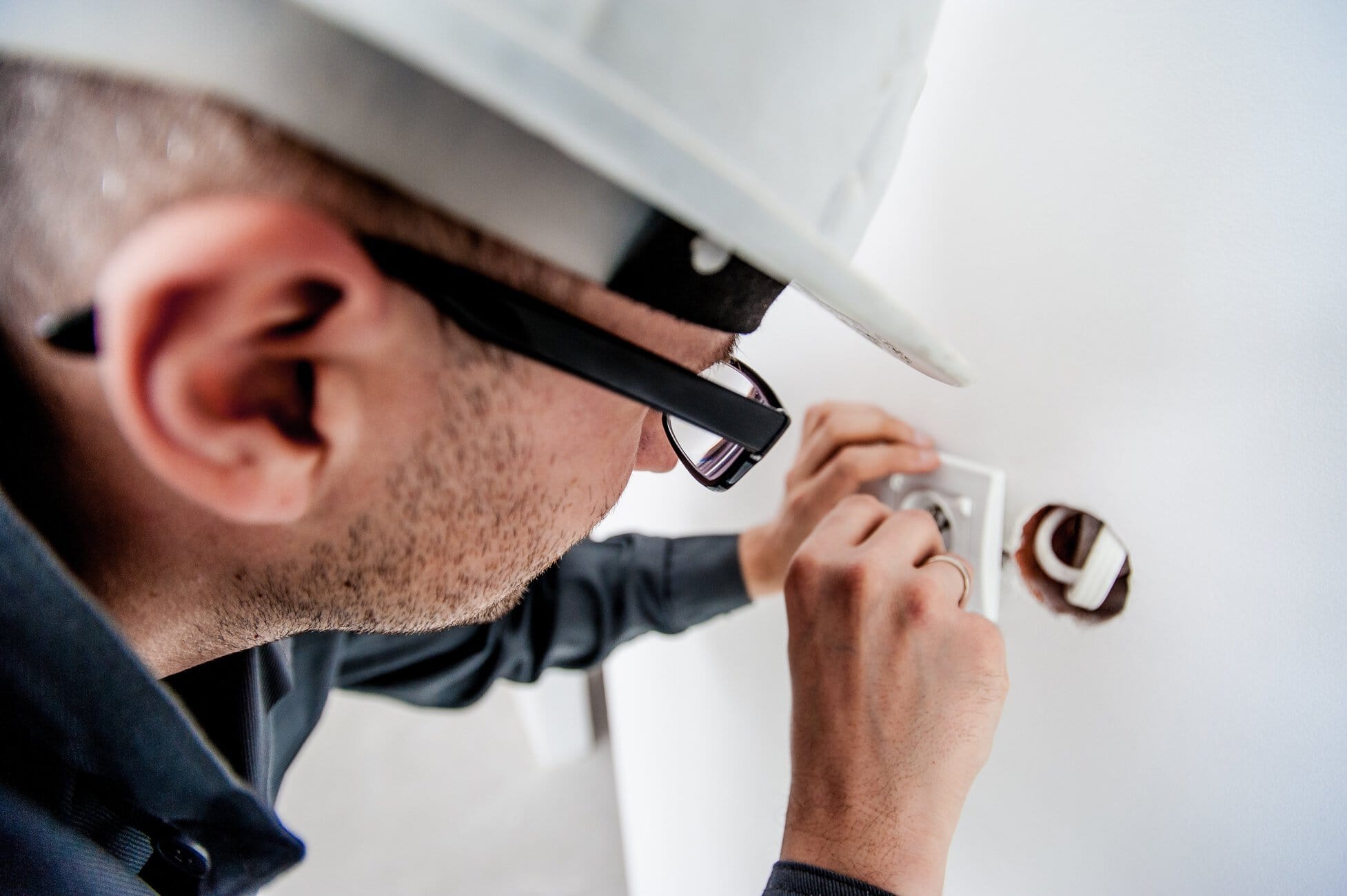 Close-up of a person wearing a white hard hat plugging a plug into a wall outlet, wiring visible nearby