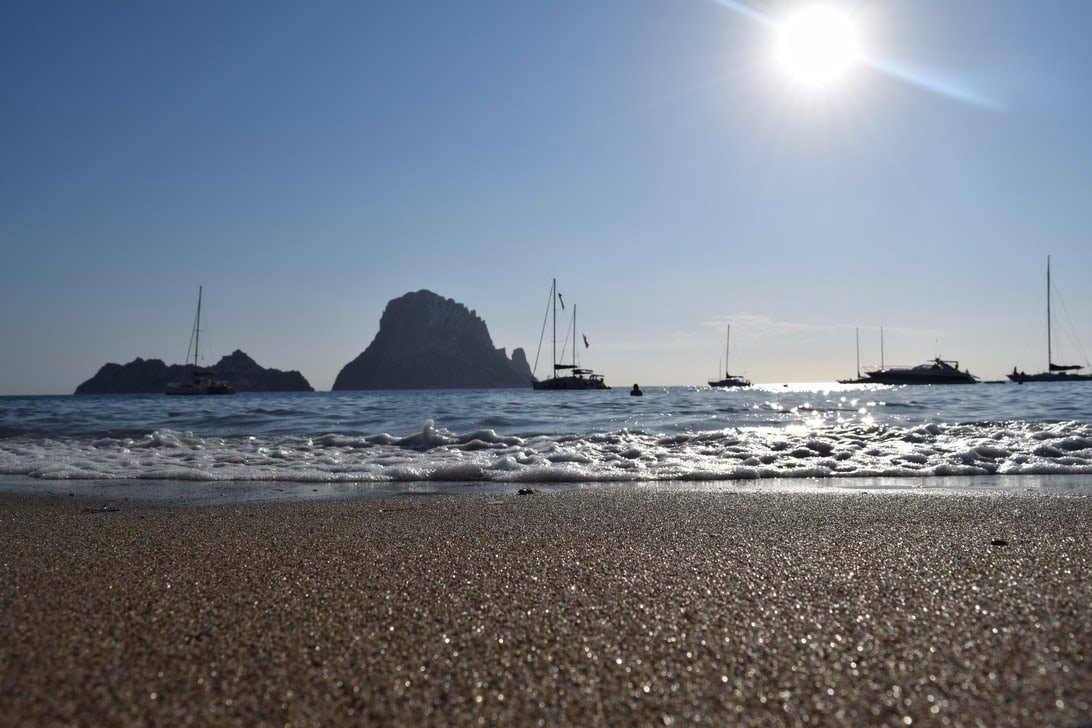Sunny beach scene with small boats anchored offshore and a rocky island in the distance under a bright sun.
