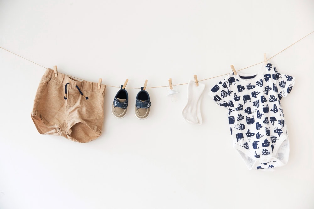 Baby clothes hanging on a line: tan shorts, small navy sandals, white socks, and a blue-printed onesie against a pale wall
