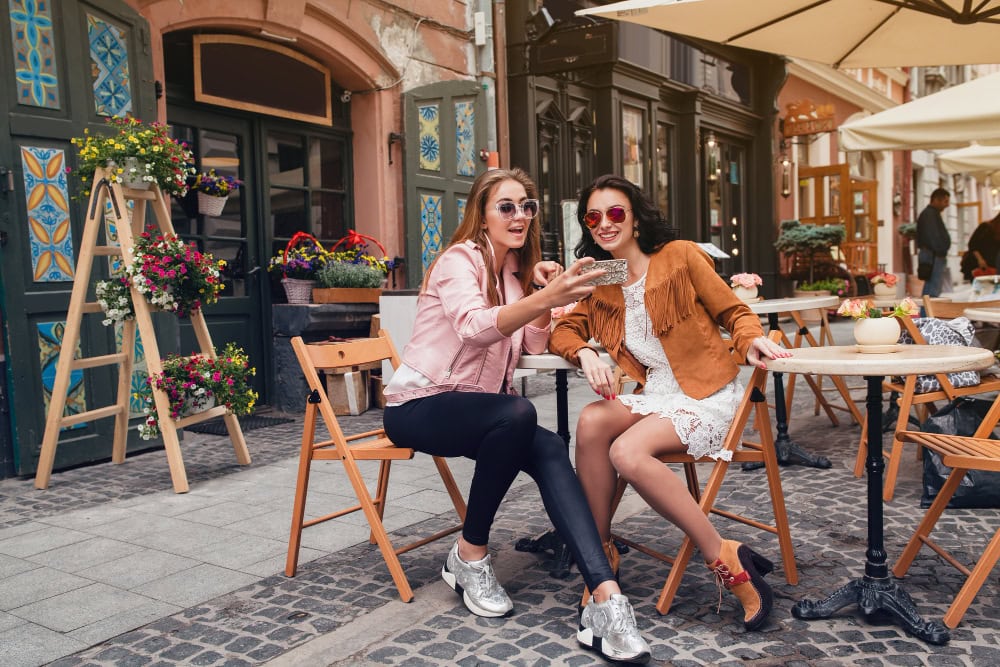 Two friends take a selfie at an outdoor cafe on a cobblestone street with flowers and umbrellas around them, smiling for the camera.