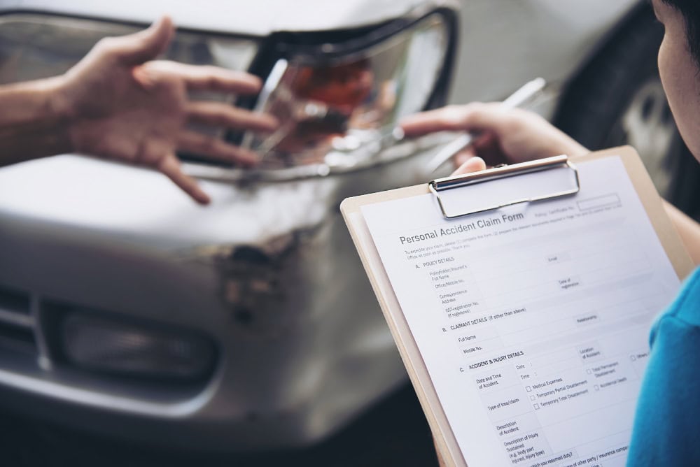 Person holds a clipboard with a Personal Accident Claim Form beside a damaged car after an accident, awaiting details from the other driver.