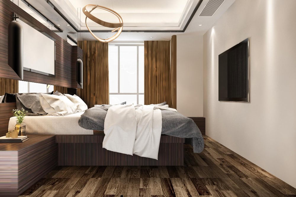 Modern hotel bedroom with a low platform bed, layered white and gray bedding, dark wood accents, a circular pendant light, and a wall-mounted TV.