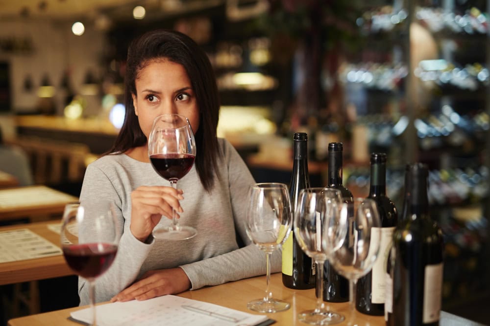 Woman at a wine bar sipping red wine, holding a stemmed glass while bottles and more glasses line the table behind her.