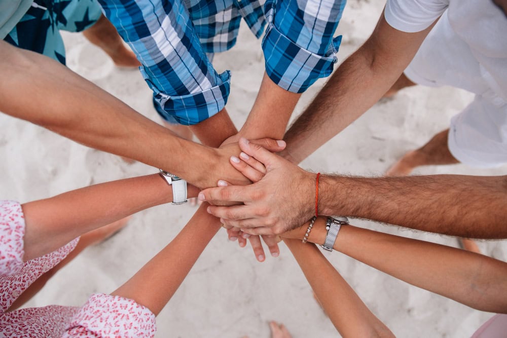Group of diverse people placing hands together in a circle on a sandy beach, symbolizing teamwork and unity.
