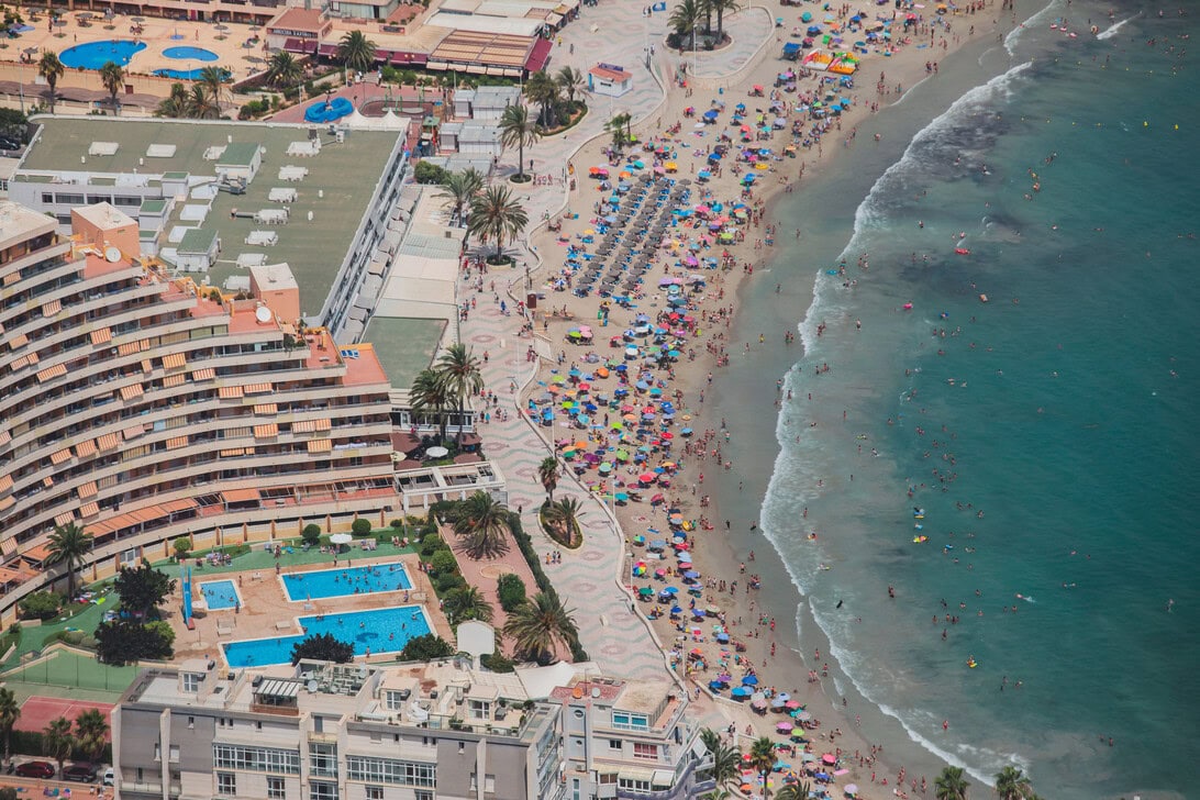 Aerial view of a crowded beach resort with colorful umbrellas along a palm-lined promenade and turquoise ocean to the right, with blue pools and buildings on the left.