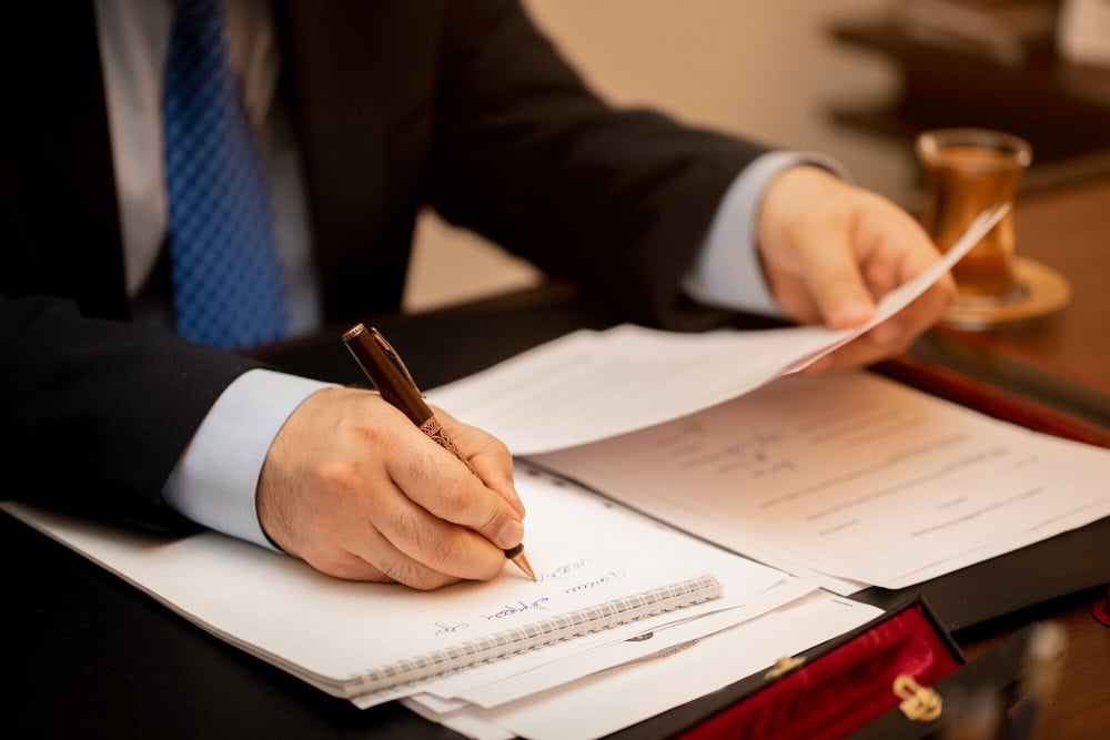 Person in a suit writes with a brown pen over documents on a desk, a cup in the background