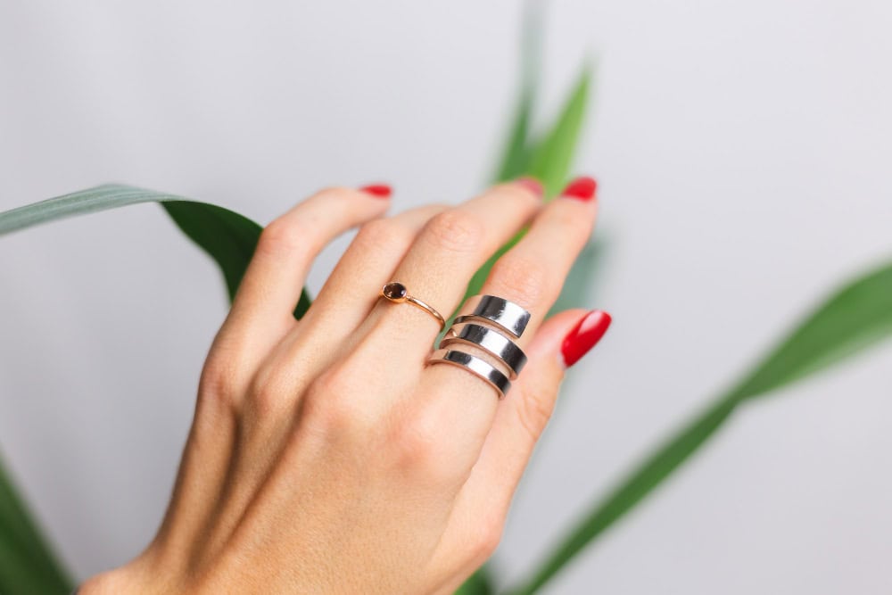 Close-up of a hand with red nail polish wearing two wide silver rings on the middle finger, with green plant leaves in the background.