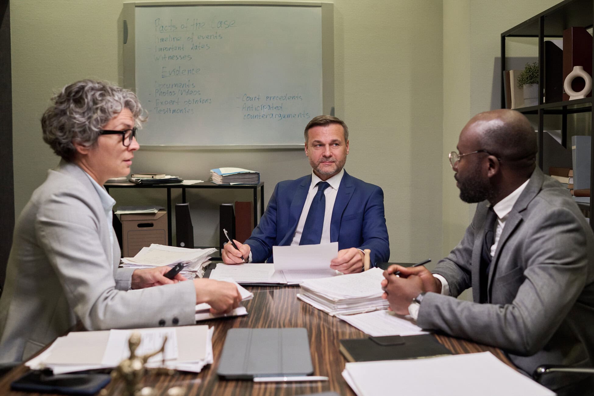 Three professionals in suits sit around a table covered with papers, reviewing documents in a meeting.