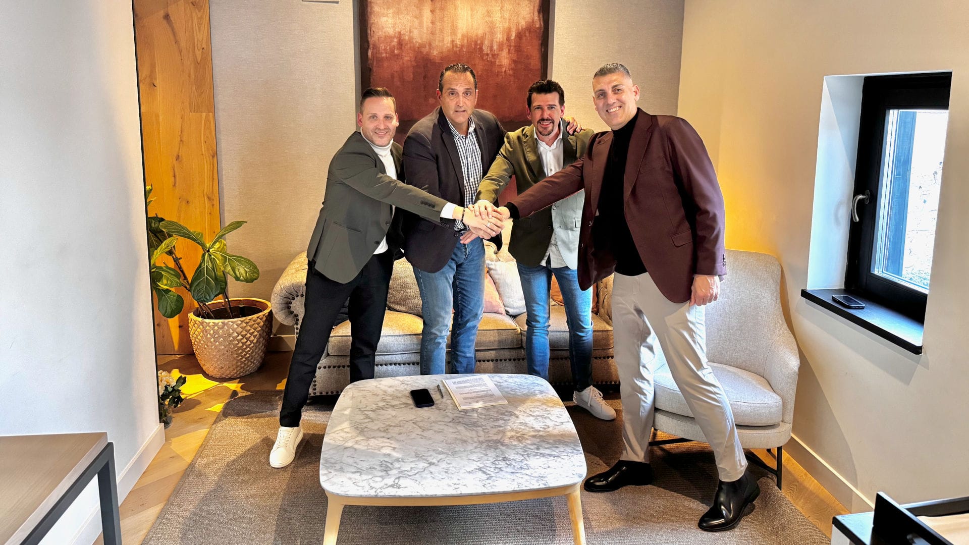 Four men in smart-casual attire stand around a marble coffee table, joining hands in a group handshake in a modern lounge setting.