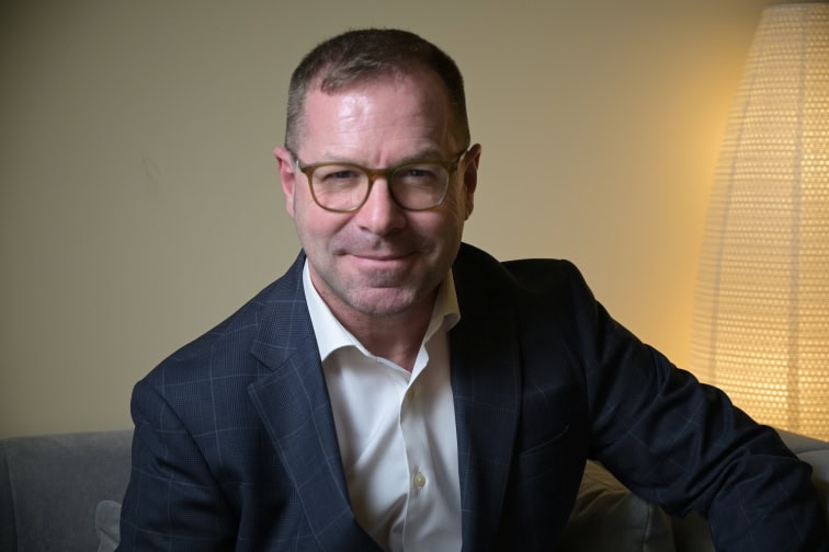 Portrait of a middle-aged man in a navy suit and white shirt sitting on a couch, smiling, wearing glasses.