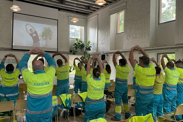 Parques workers in yellow and blue uniforms stretch arms overhead during a training session in a bright room with a projector showing a fitness instructor on screen.