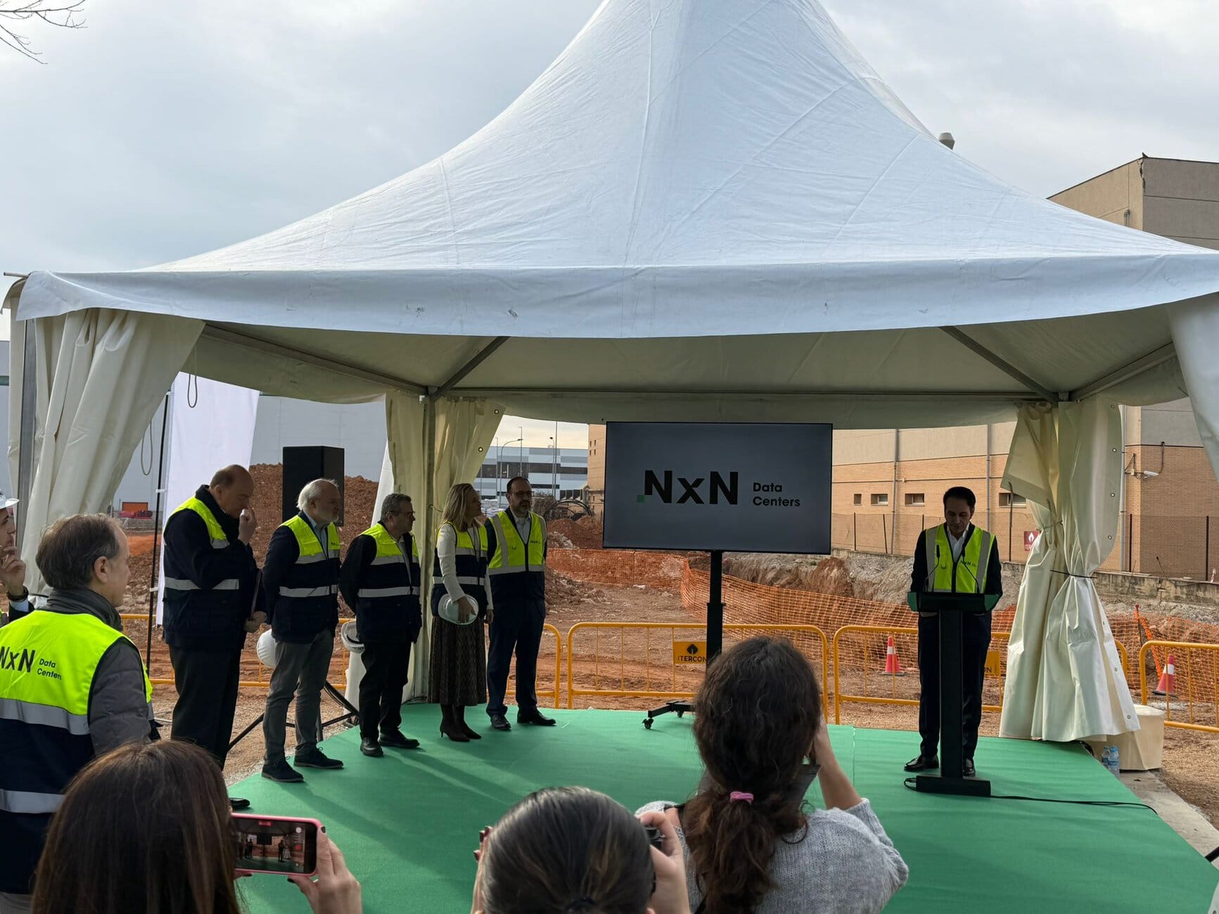 Group of people in high-visibility vests under a white event tent, listening to a speaker with a screen that reads 'NxN Data Centers'.