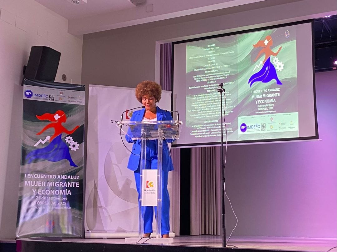 Female speaker at a podium on a stage during a conference, with banners and a projection screen showing the event poster read 'Mujer Migrante y Economía'.