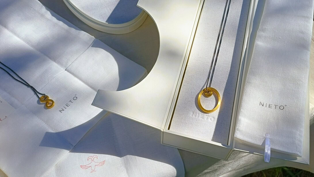 Gold necklace with two interlocking circular pendants displayed on white fabric with Nieto branding and boxes nearby.