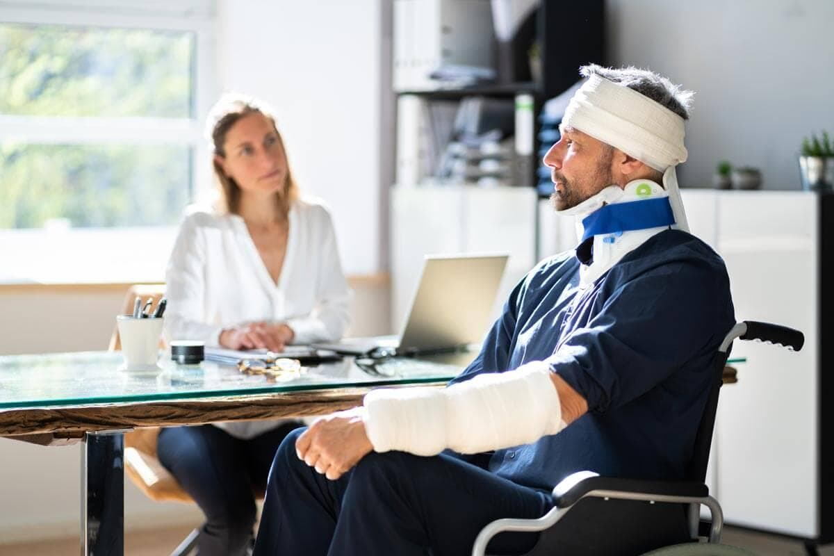Man with a head bandage and arm in a cast sits in a wheelchair across a glass desk, talking with a woman in an office setting.