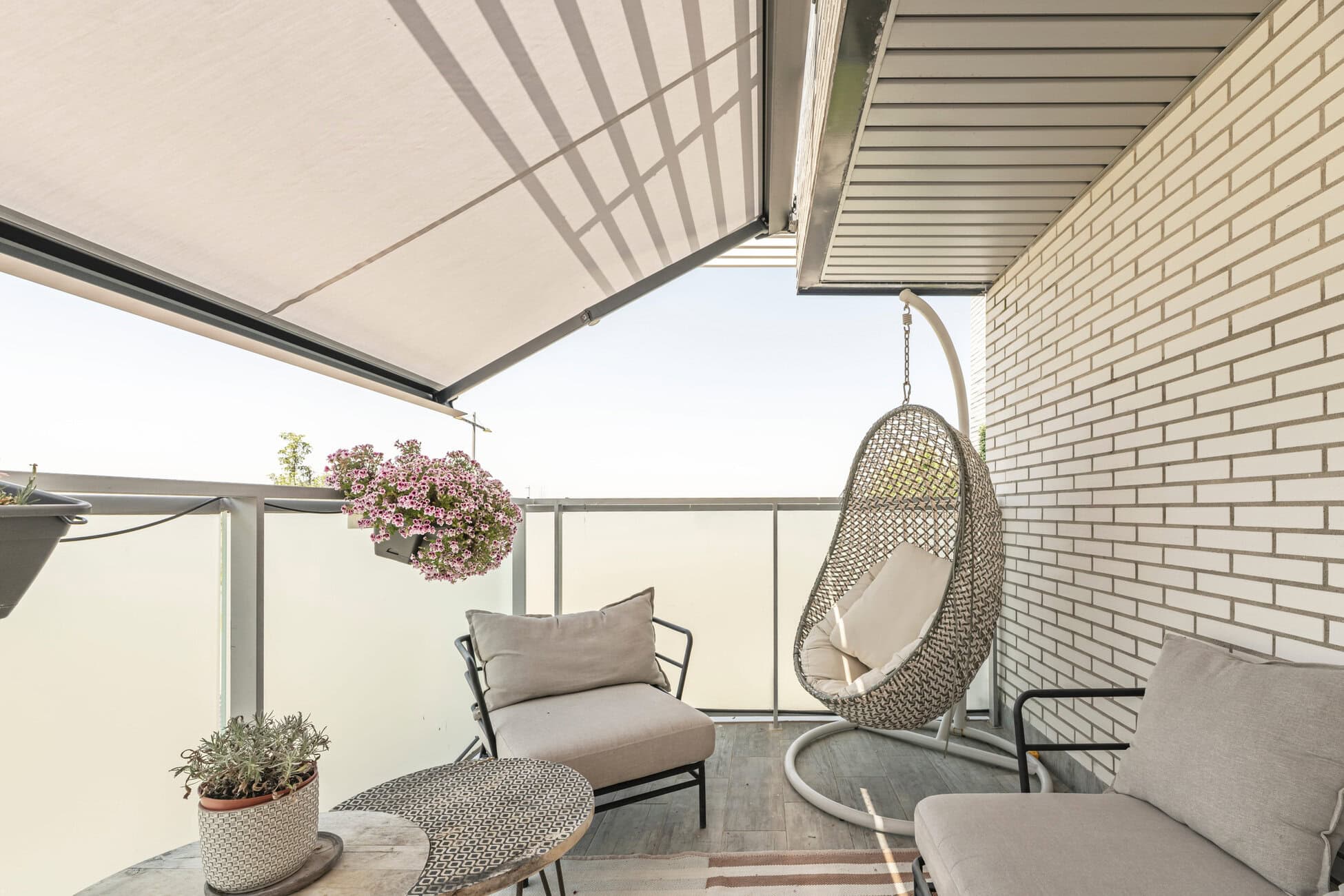 Cozy outdoor balcony with beige cushioned seating, a hanging wicker egg chair, and potted flowers beside a glass railing.