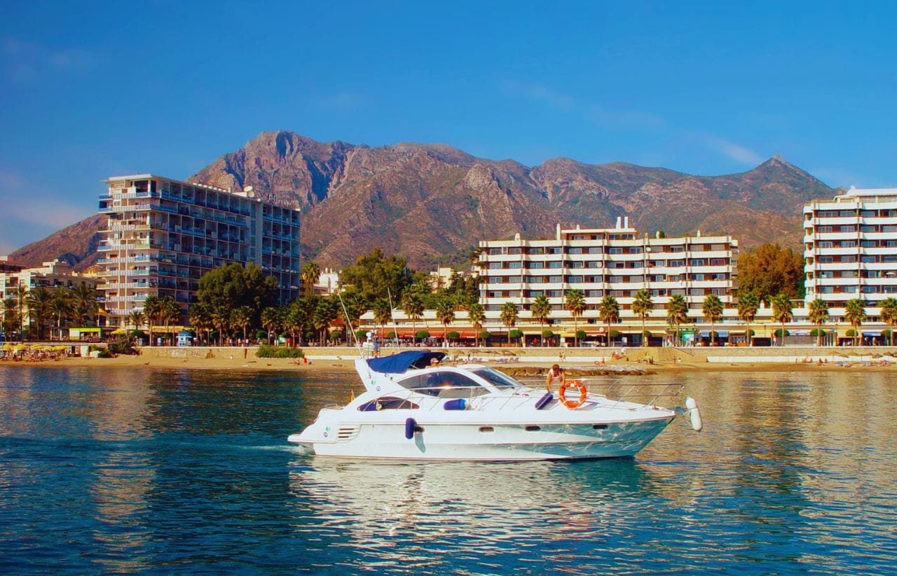 White motor yacht sailing in calm blue water with a beachfront resort and palm trees, and rugged mountains in the background