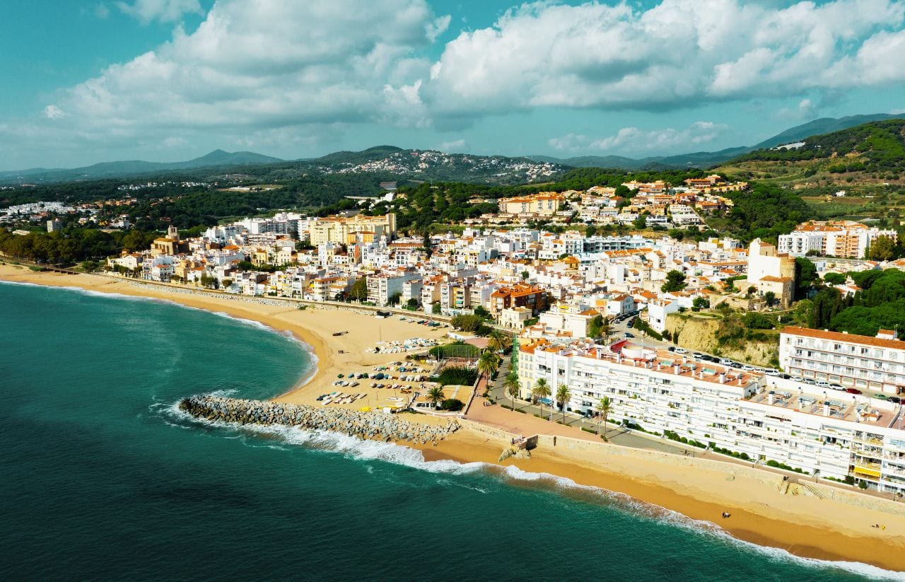 Aerial view of a Mediterranean coastal town with white and pastel buildings along a curved golden beach and turquoise sea.