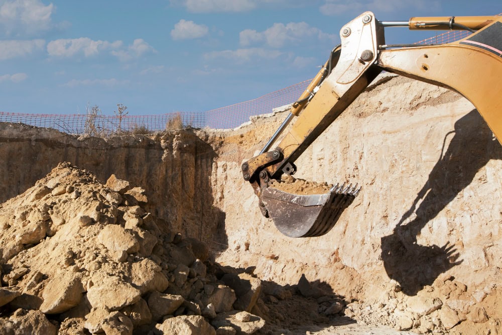 Excavator bucket scooping soil from a deep trench at a construction site; dirt pile in foreground and blue sky above.