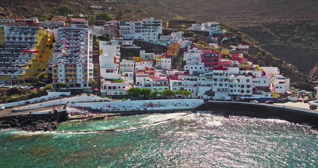 Colorful hillside town by the sea with white breakwater and turquoise water along a rocky shore, overlooking a narrow coastal road.