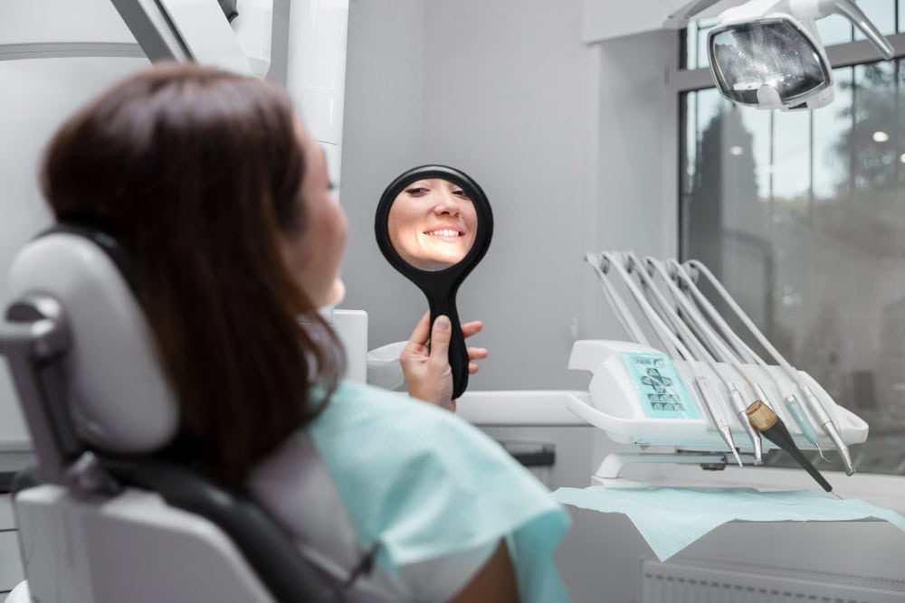 Patient in a dental chair looks into a handheld mirror and smiles, with dental tools nearby in the clinic.