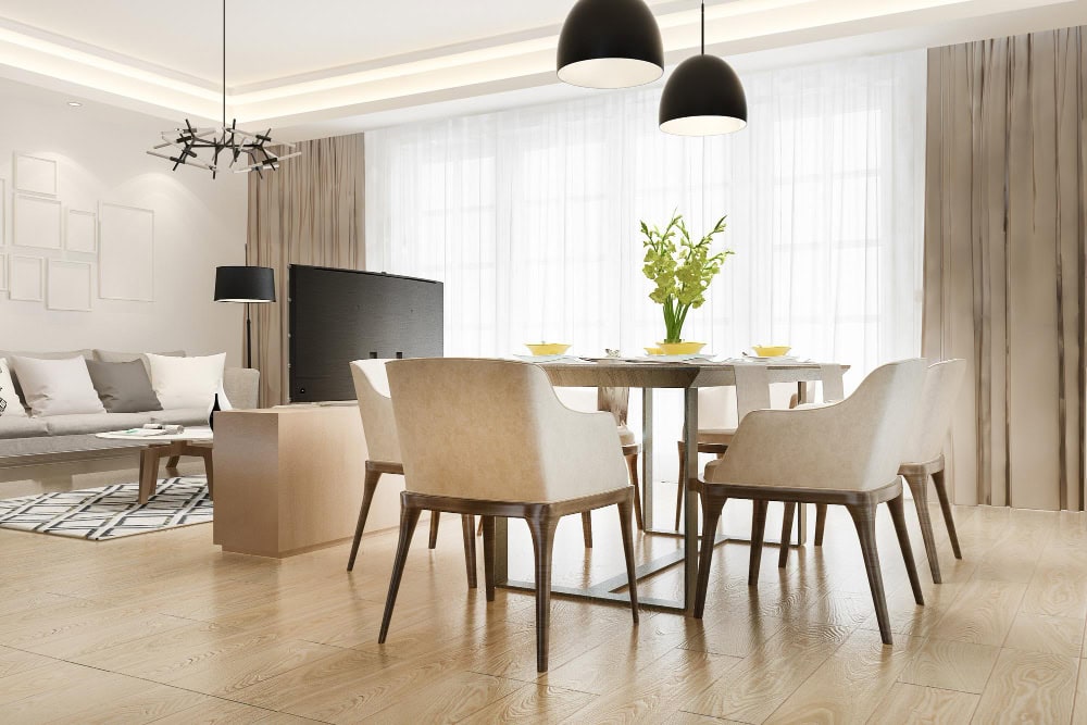 Open-plan living and dining room with a beige dining table and six matching chairs, a gray sofa, and large windows with sheer curtains.