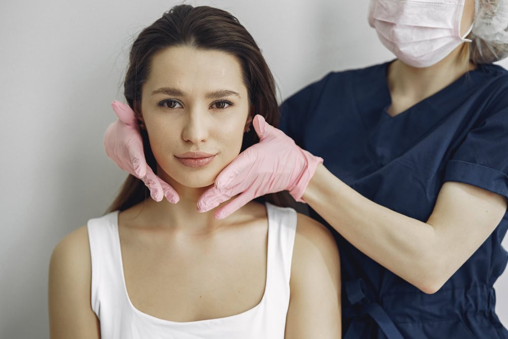 Healthcare professional in blue scrubs and pink gloves holds a woman's jawline during a checkup.
