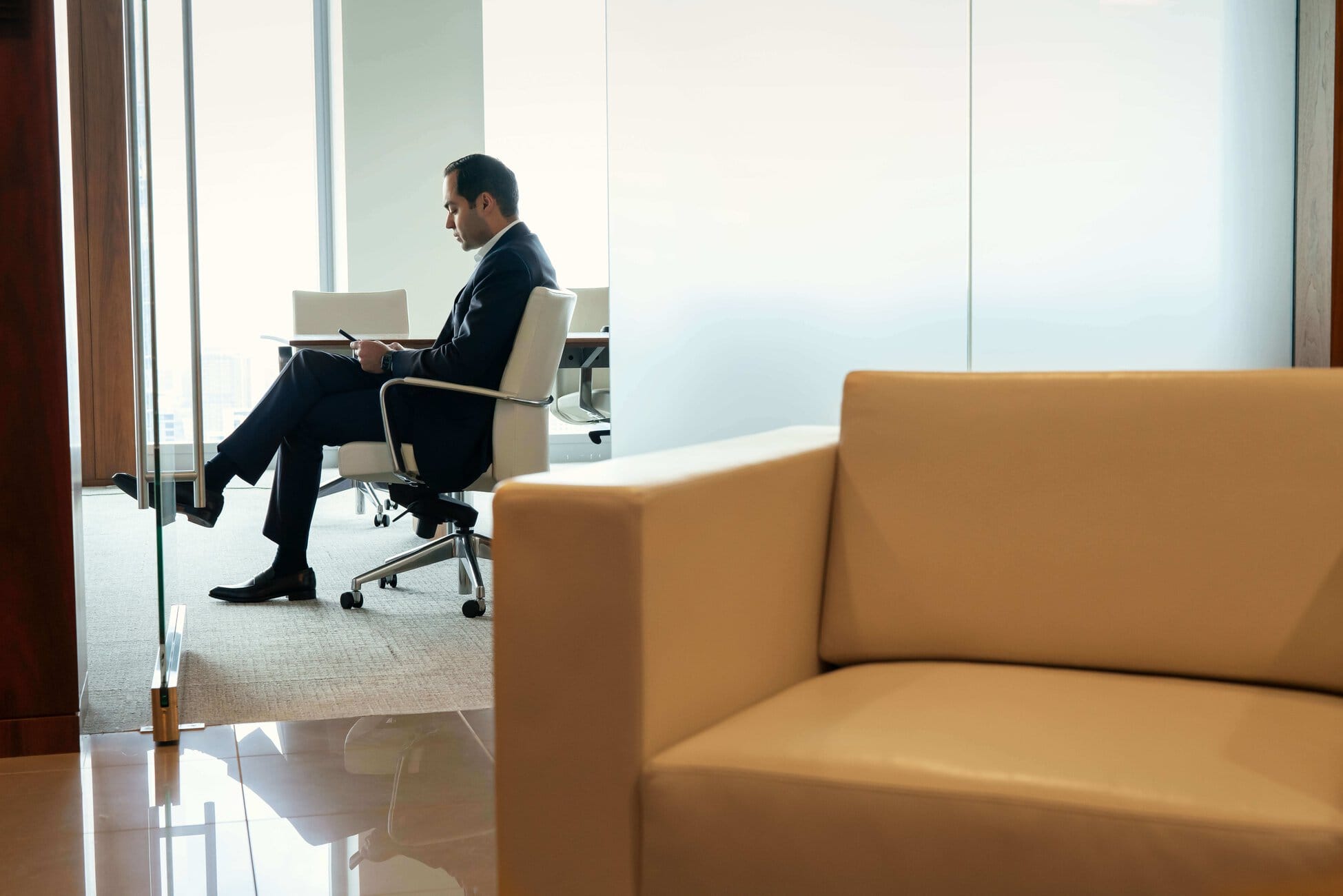 Man in a dark suit sits in an office chair at a glass desk, looking at a smartphone, with a beige sofa in the foreground.