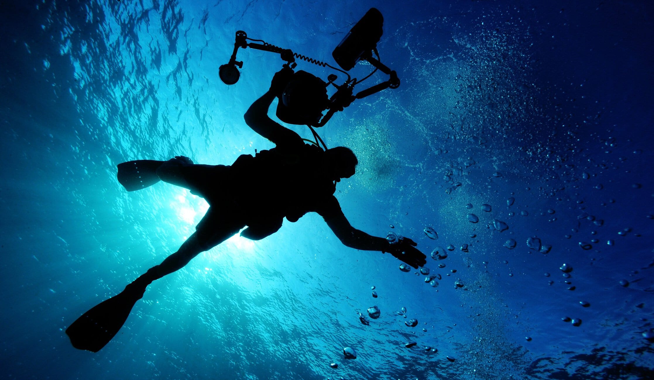 Two divers underwater, one holding a camera rig, silhouetted against the blue ocean and bubbles.