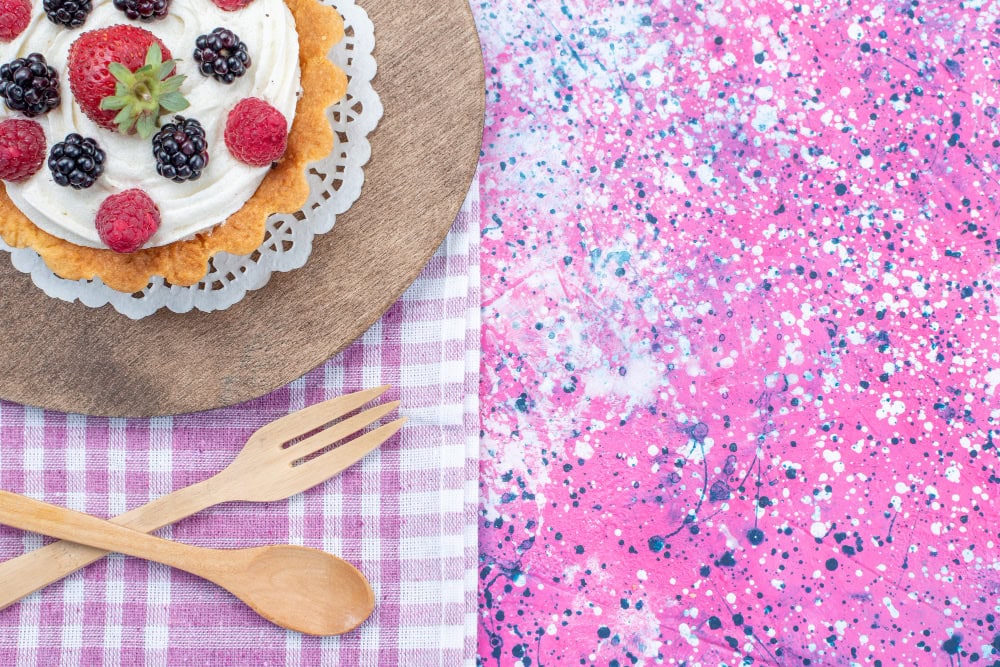 Berry-topped tart on a wooden board with a lace doily, beside a pink speckled background and a pink gingham cloth with wooden cutlery.