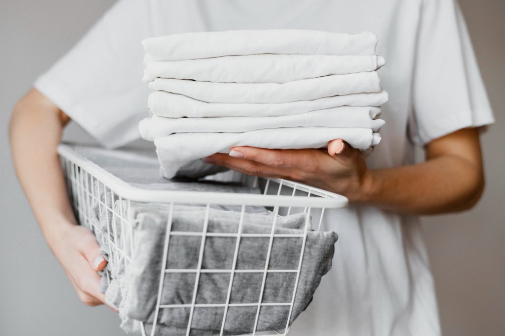 Person in a white shirt holding a laundry basket filled with folded white towels and a grey cloth.